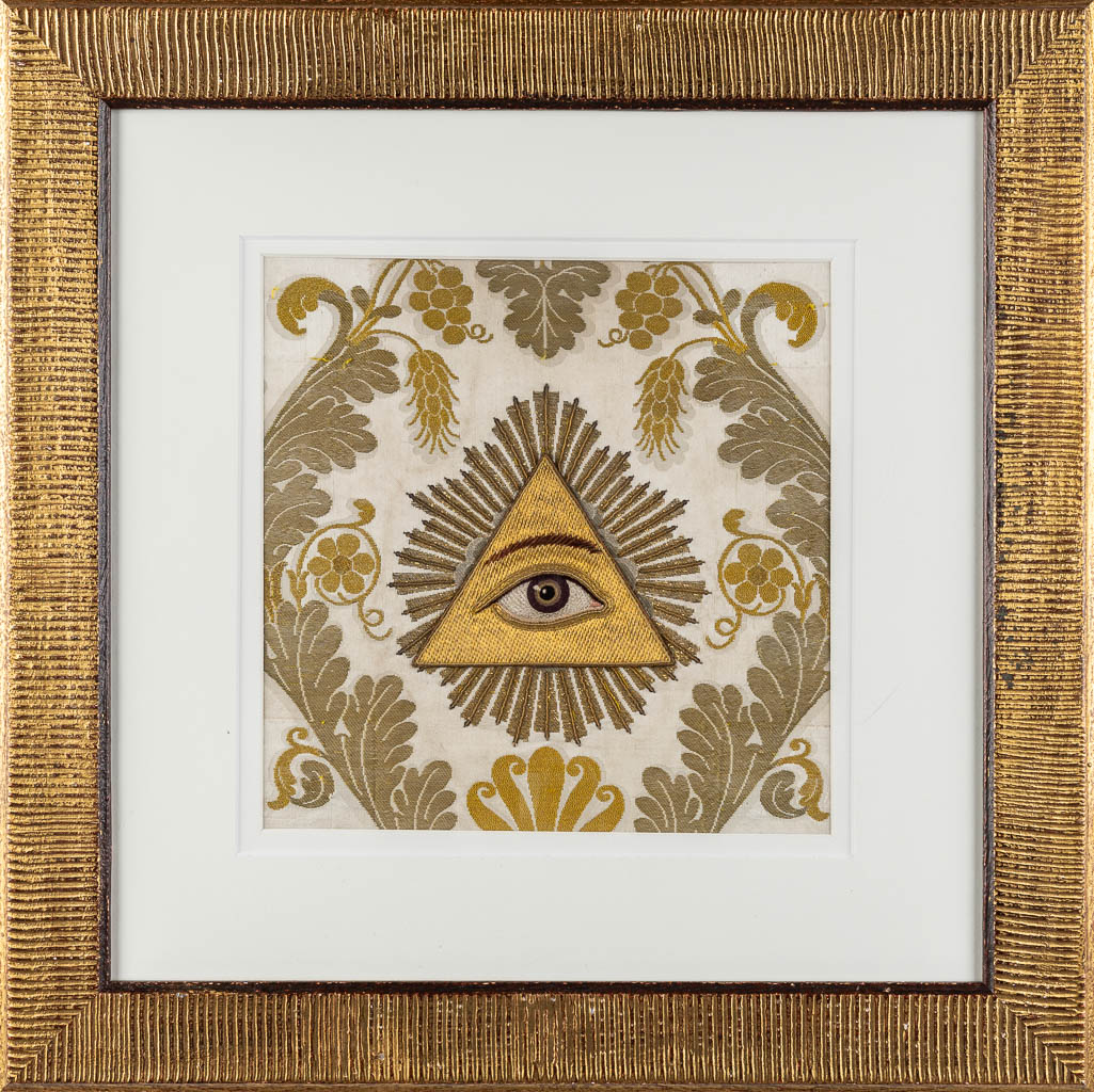 Een ingekaderd 'Alziend Oog', goudbrokaat borduurwerk. (W:29,5 x H:29,5 cm)