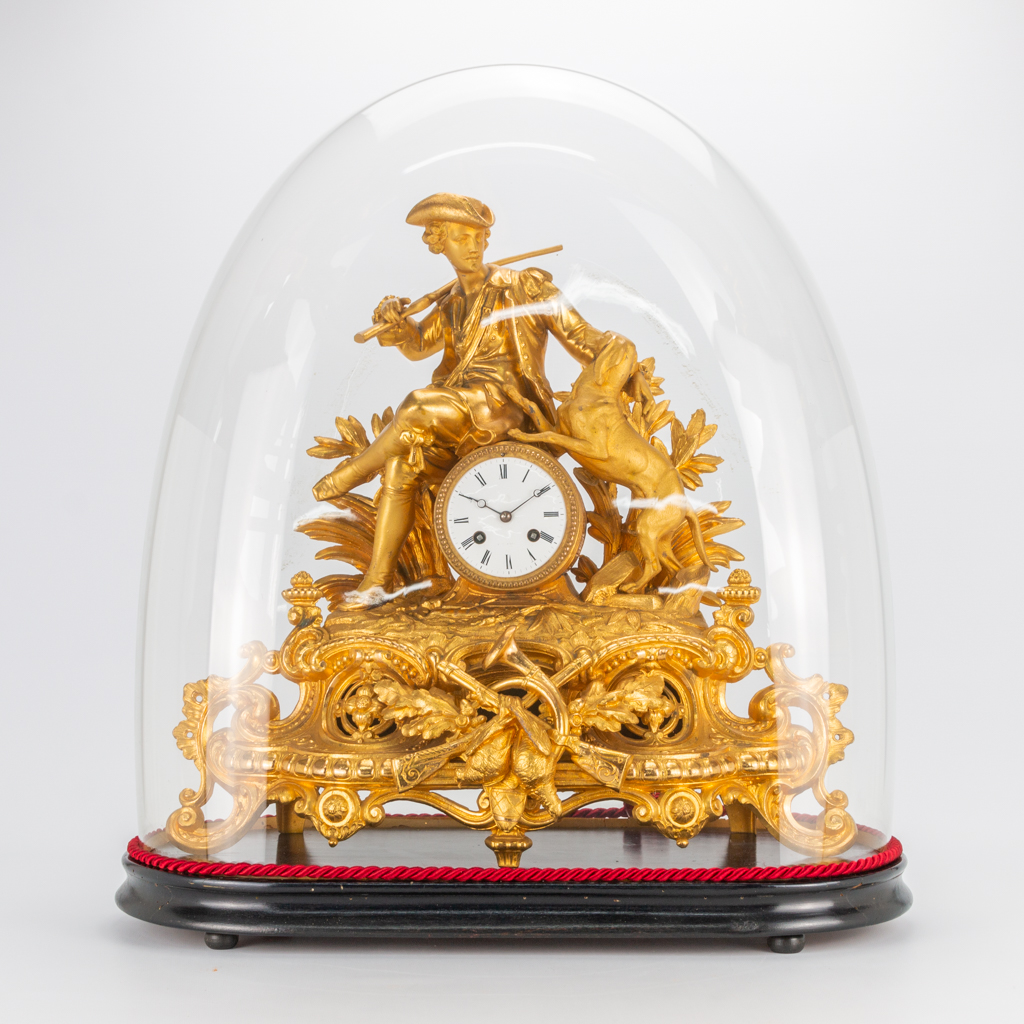 A mantle clock, with a hunter figurine and his dog, hunting trophies. Under a glass dome. 19th century. 