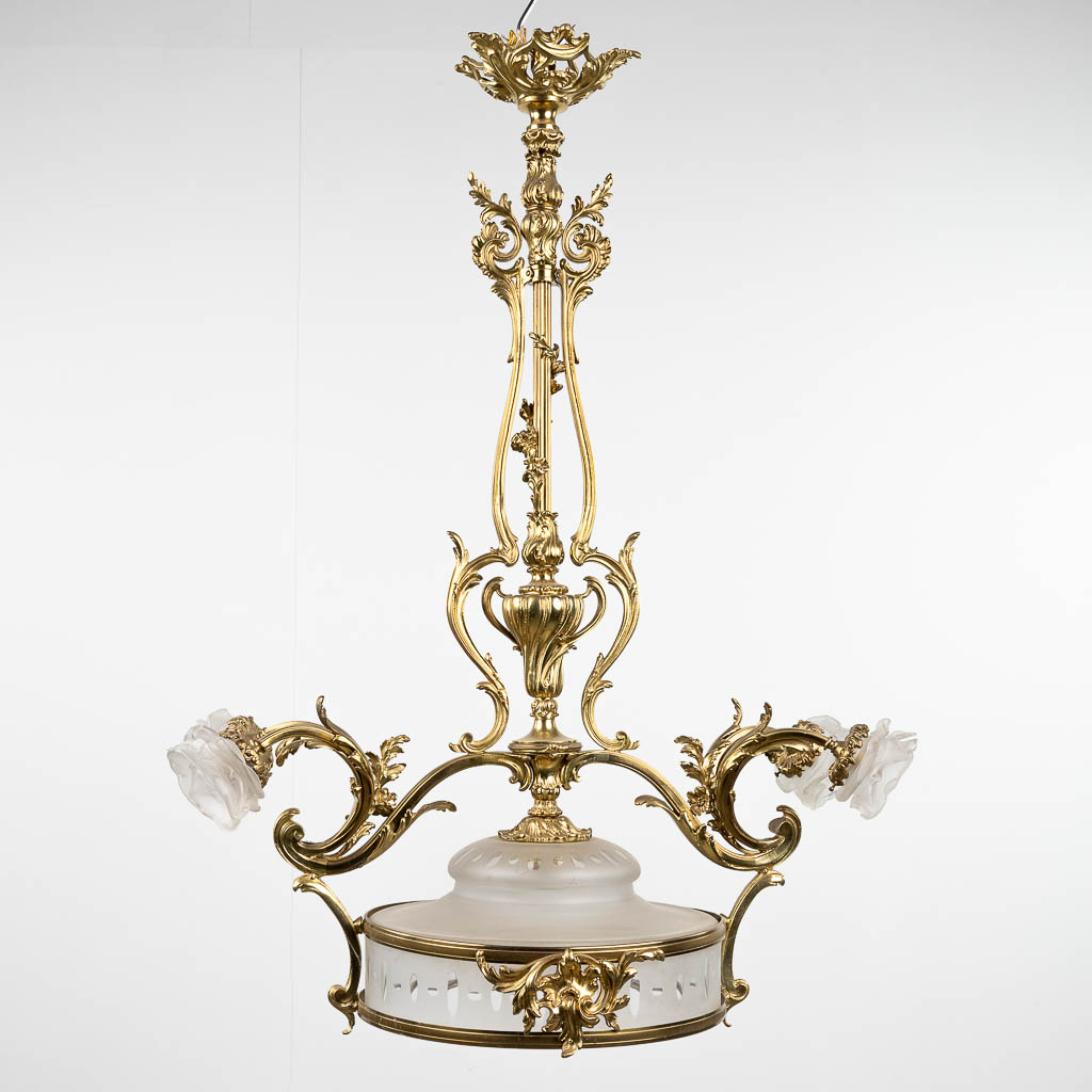 Een luchter, brons in Lodewijk XV stijl, afgewerkt met glazen lampenkappen en schaal. (L: 38 x W: 70 x H: 100 cm)