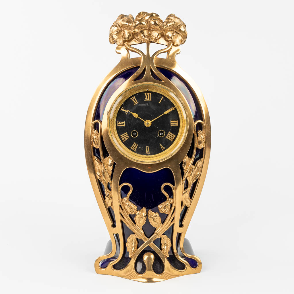 A table- or mantle clock made of porcelain mounted with bronze in art nouveau style.  (L:13 x W:19 x H:38 cm)