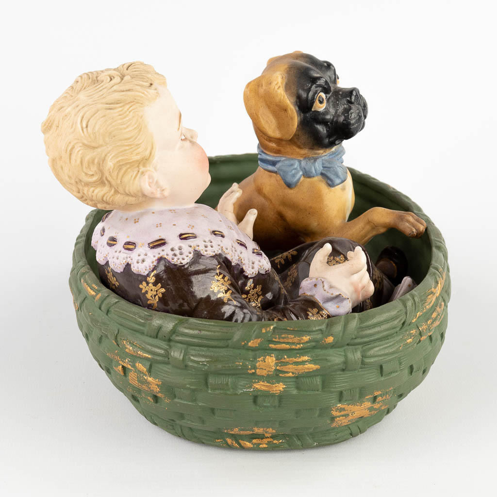 Een kind met een hond in een mand, polychrome biscuit porselein. Circa 1900. (D:18 x W:24 x H:16 cm)
