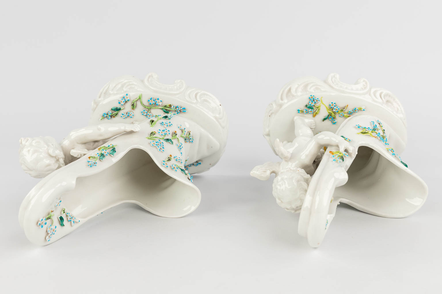 Sitzendorf, een paar bloemenvazen gemaakt uit porselein, in de vorm van 2 putti met een schoen. (L: 12,5 x W: 16 x H: 25 cm)