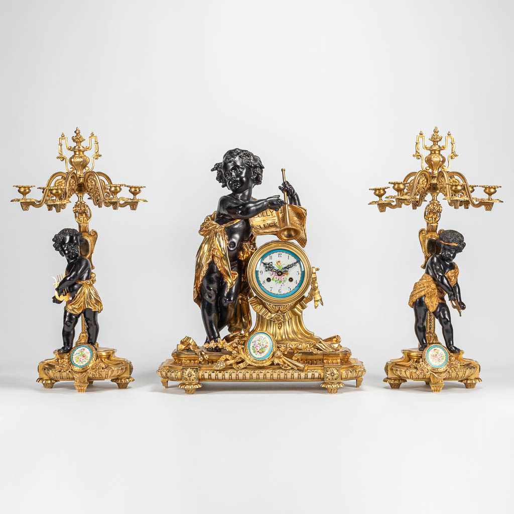 Een driedelige schouwgarnituur met bronzen klok en kandelaars, versierd met putti en handgeschilderde porseleinen plaquettes.