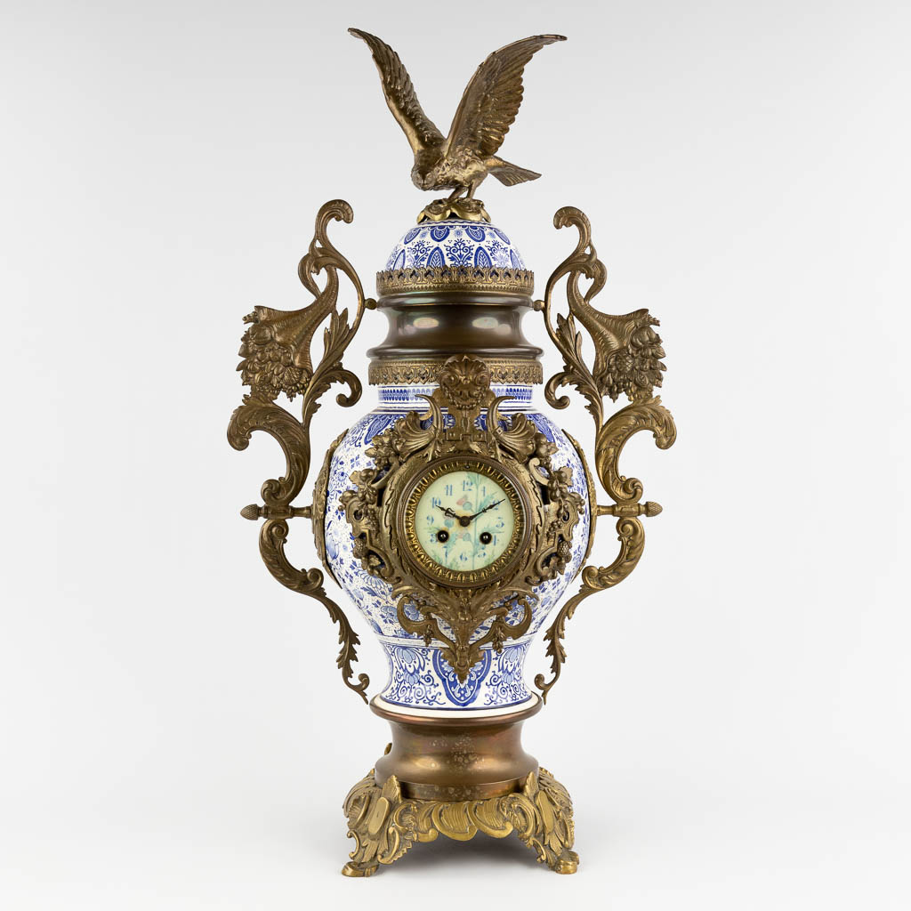 A mantle clock, Delfts faience mounted with bronze and an eagle figurine. Circa 1900. (D:25 x W:44 x H:81 cm)