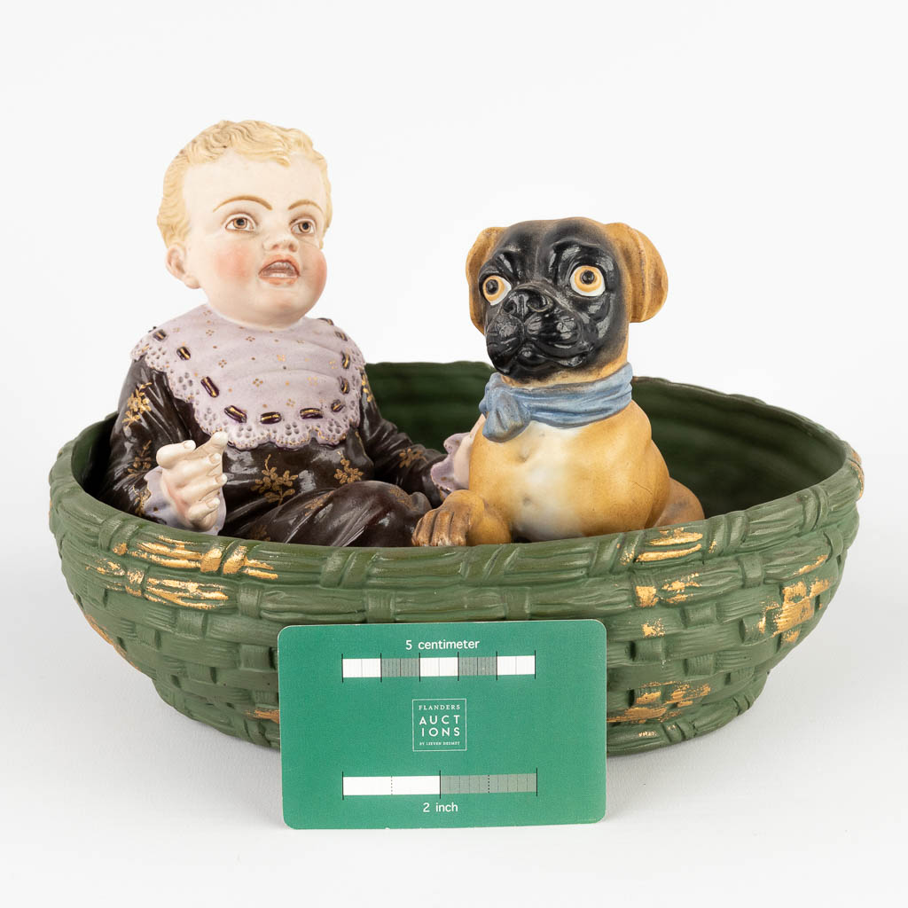 Een kind met een hond in een mand, polychrome biscuit porselein. Circa 1900. (D:18 x W:24 x H:16 cm)